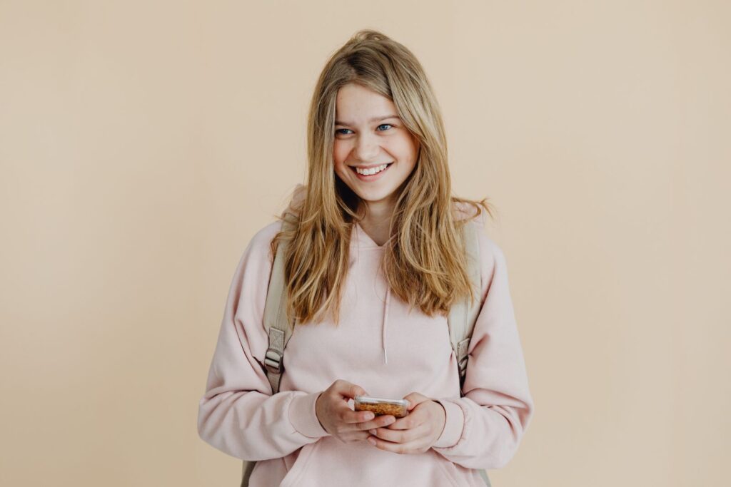 A cheerful teenager with blonde hair wearing a pink sweater and using a smartphone indoors.