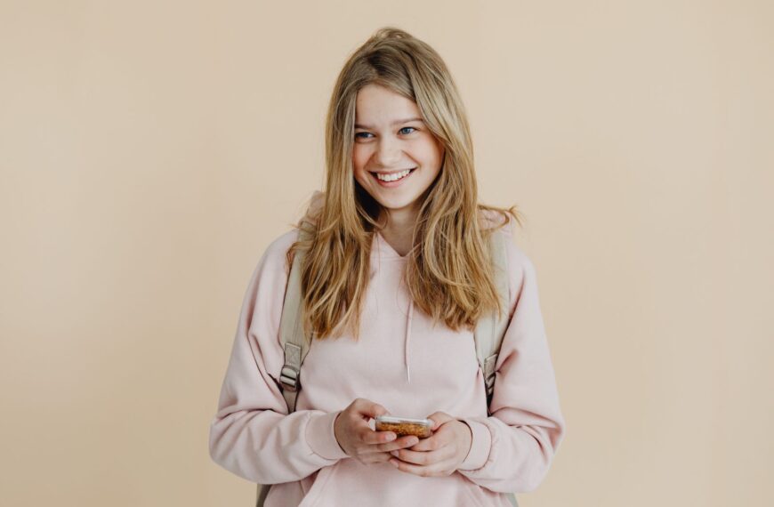 A cheerful teenager with blonde hair wearing a pink sweater and using a smartphone indoors.