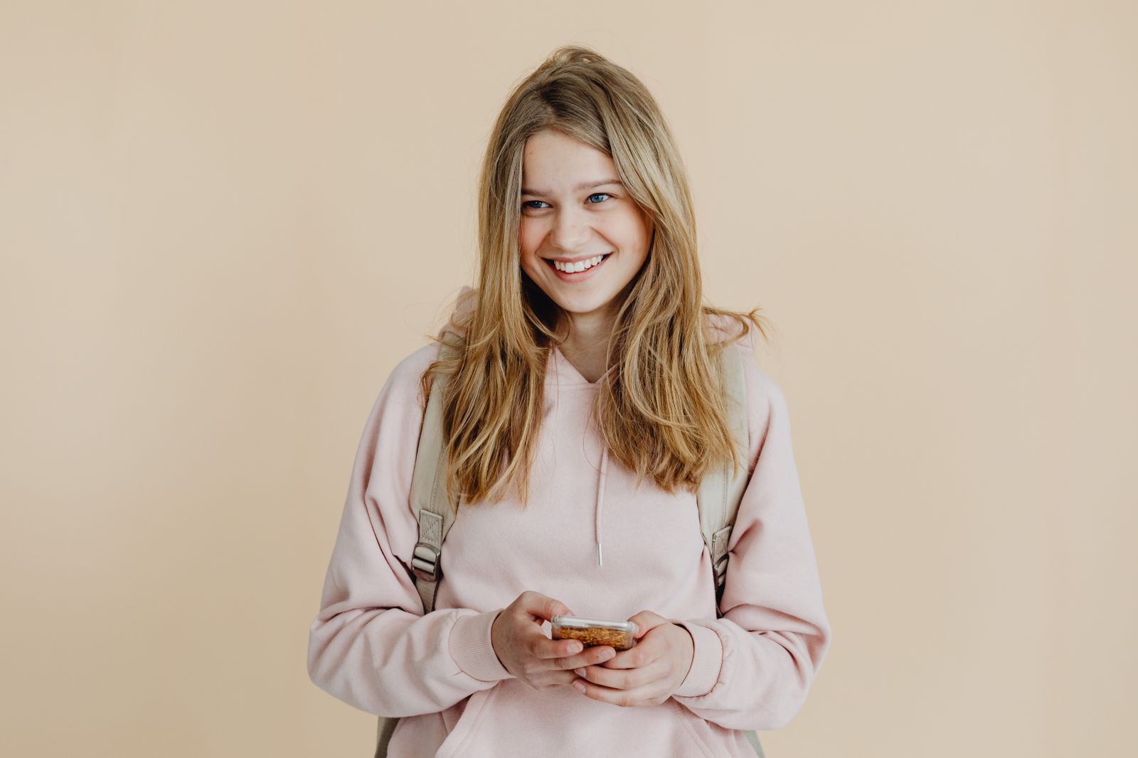A cheerful teenager with blonde hair wearing a pink sweater and using a smartphone indoors.