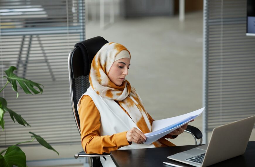 Middle Eastern businesswoman wearing a hijab reading documents at her office desk with a laptop.