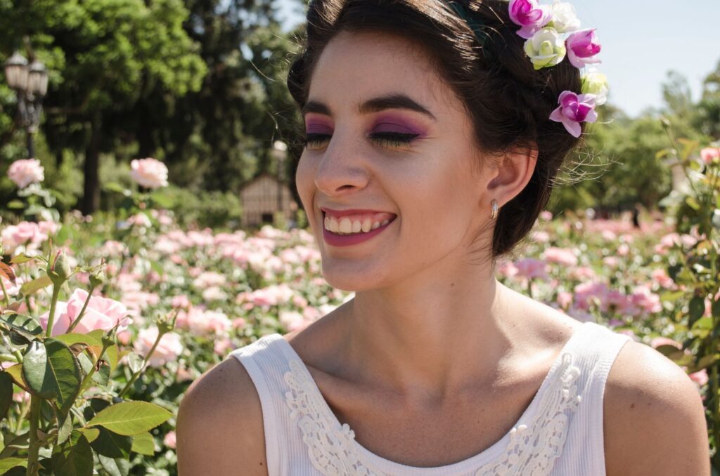 A young woman wearing a flower crown smiles amidst blooming roses in an outdoor garden.