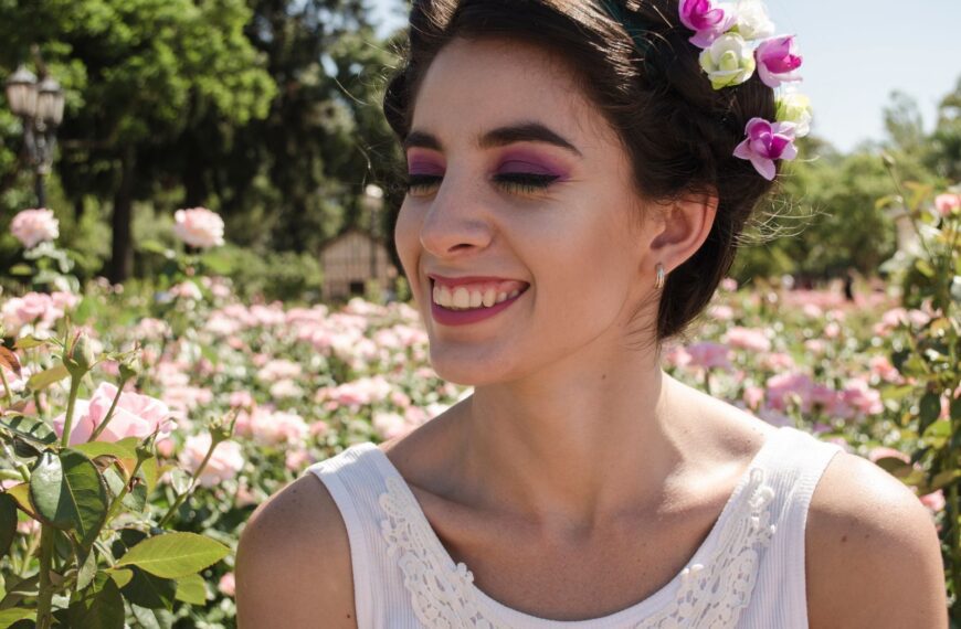 A young woman wearing a flower crown smiles amidst blooming roses in an outdoor garden.
