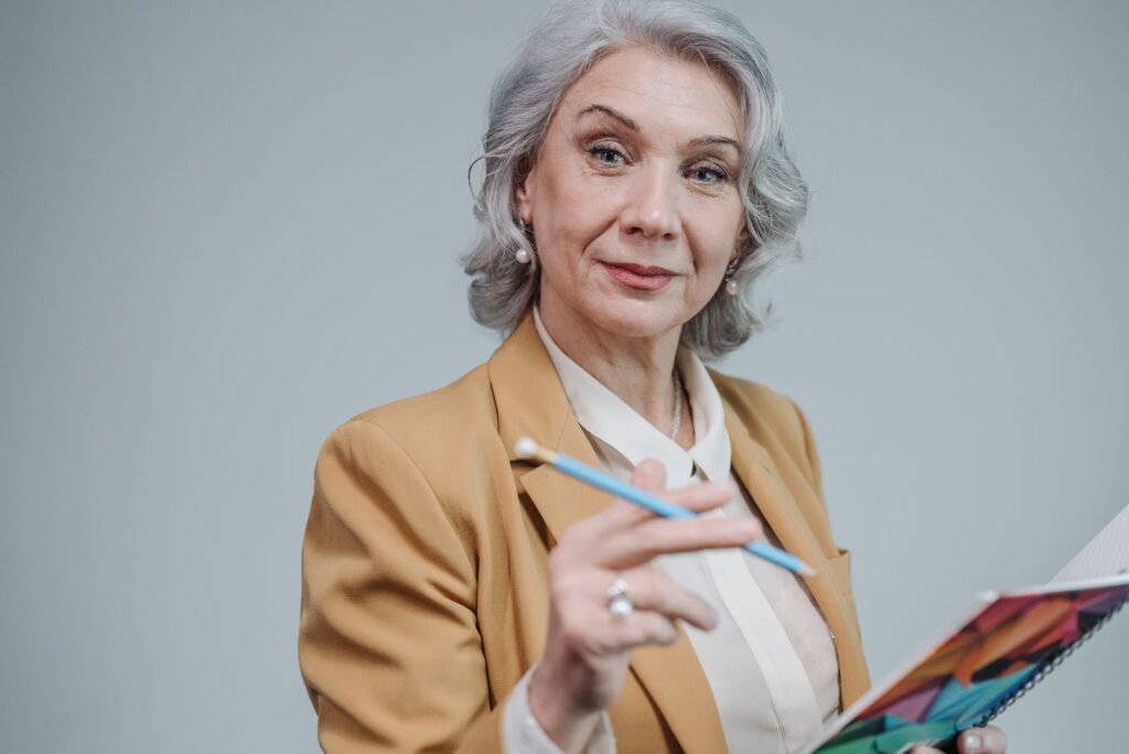 Portrait of a senior woman in a brown blazer holding a notebook and smiling warmly.