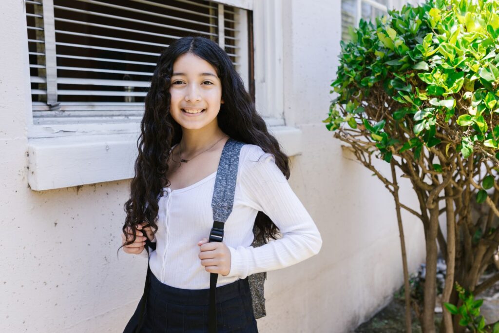A young girl with curly hair smiling at the camera, wearing a white shirt and backpack outdoors.