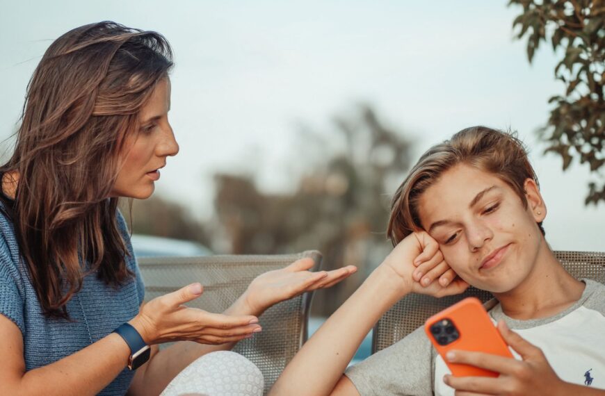 A mother engaging with her teenage son holding a smartphone outdoors.