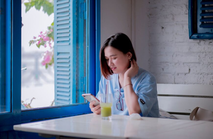 A young woman in a light blue shirt checks her smartphone while enjoying a drink by a window in a quaint cafe.
