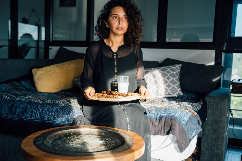 A woman in a black dress sits on a couch holding a tray of dates and milk, symbolizing iftar indoors.