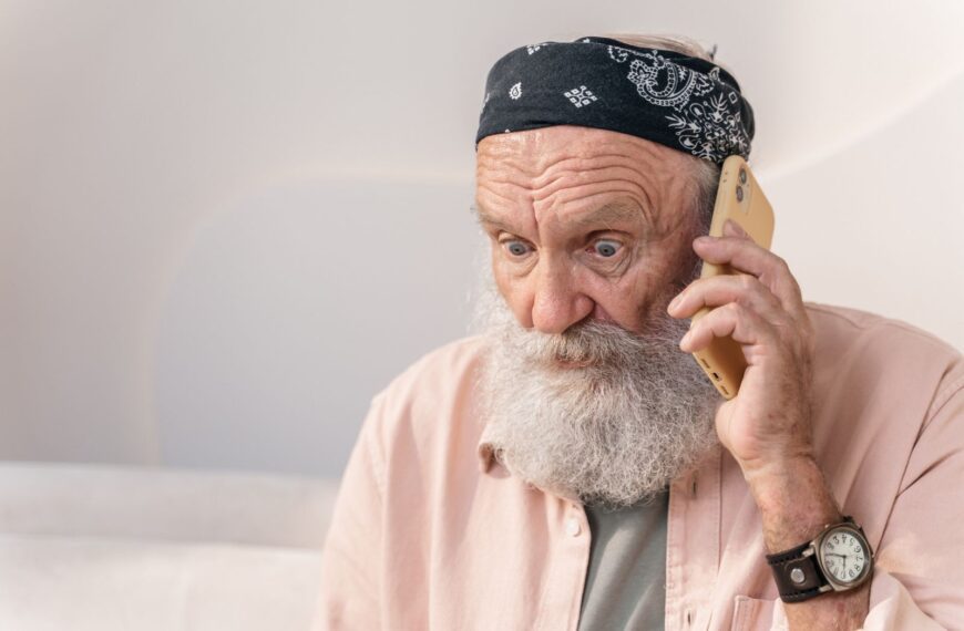 An elderly man with a beard looks surprised while on the phone indoors.