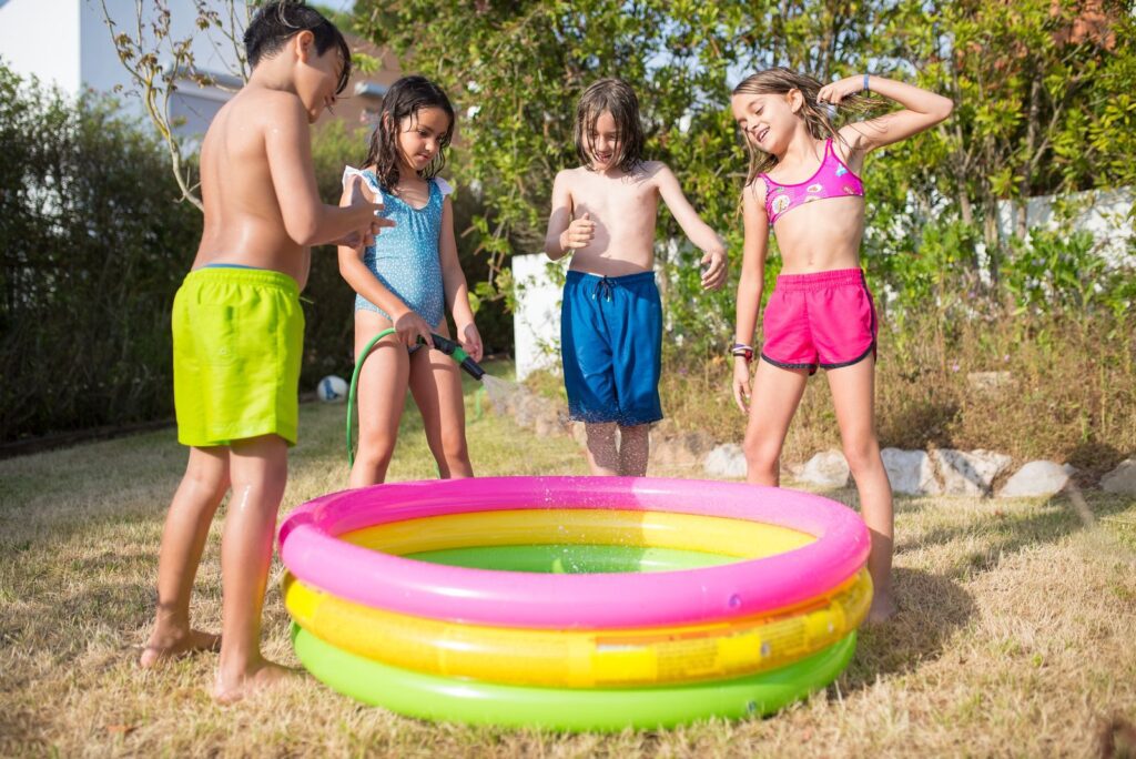 Four kids playing with a garden hose in a colorful kiddie pool on a sunny summer day.