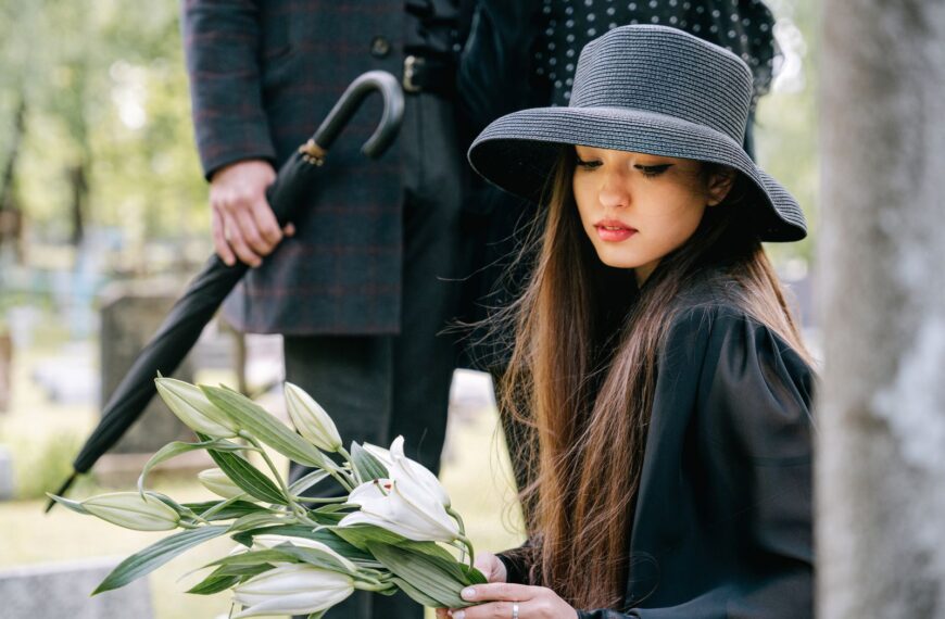 A woman in a black hat holds lilies, mourning at a cemetery.