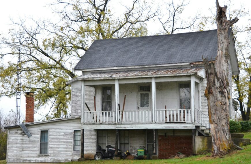 Old farmhouse with peeling paint and a large tree.