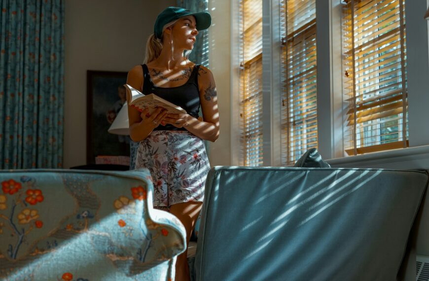 woman holding book while standing beside window blinds