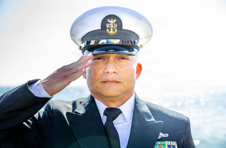 A man in a us navy uniform salutes outdoors.