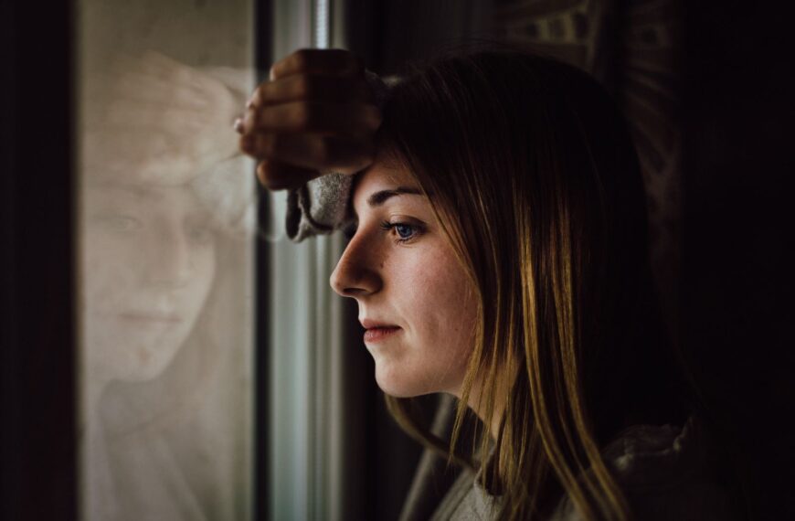 A contemplative young woman looks out a window with a reflective expression, indoors.