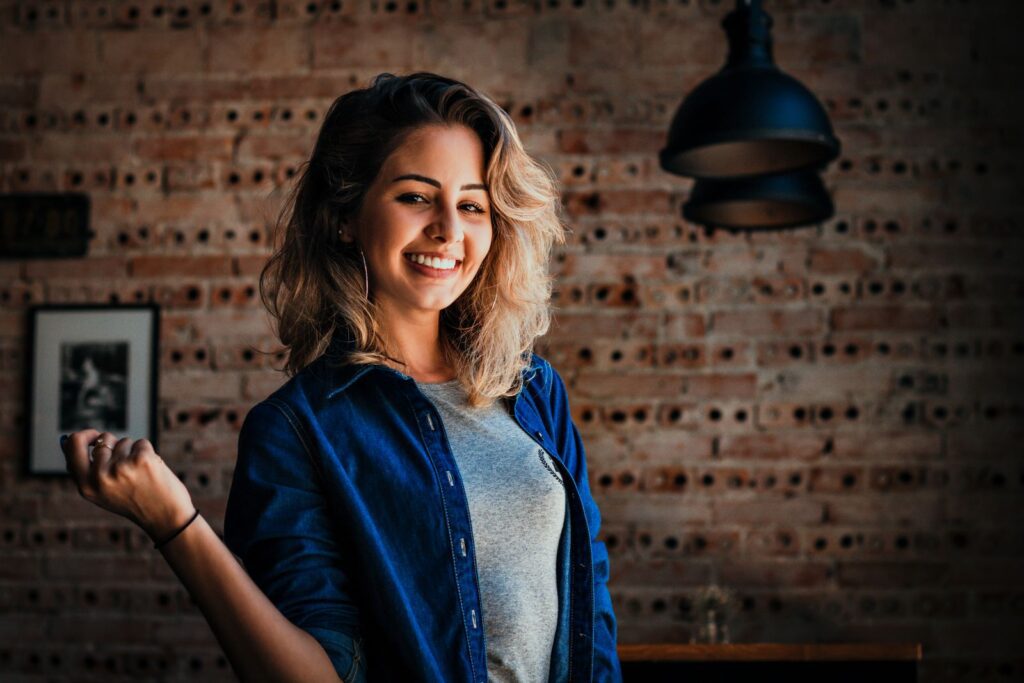 Portrait of a smiling woman in denim jacket against a brick wall indoors.