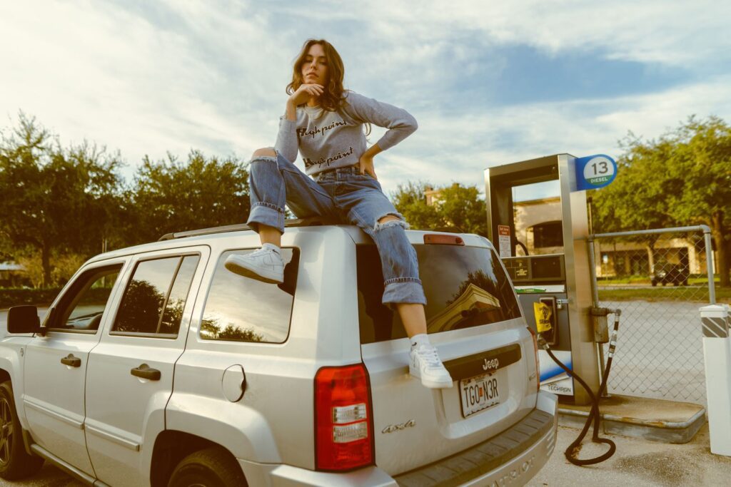 Stylish woman in casual outfit poses atop an SUV at a gas station, exuding confidence and modern fashion.