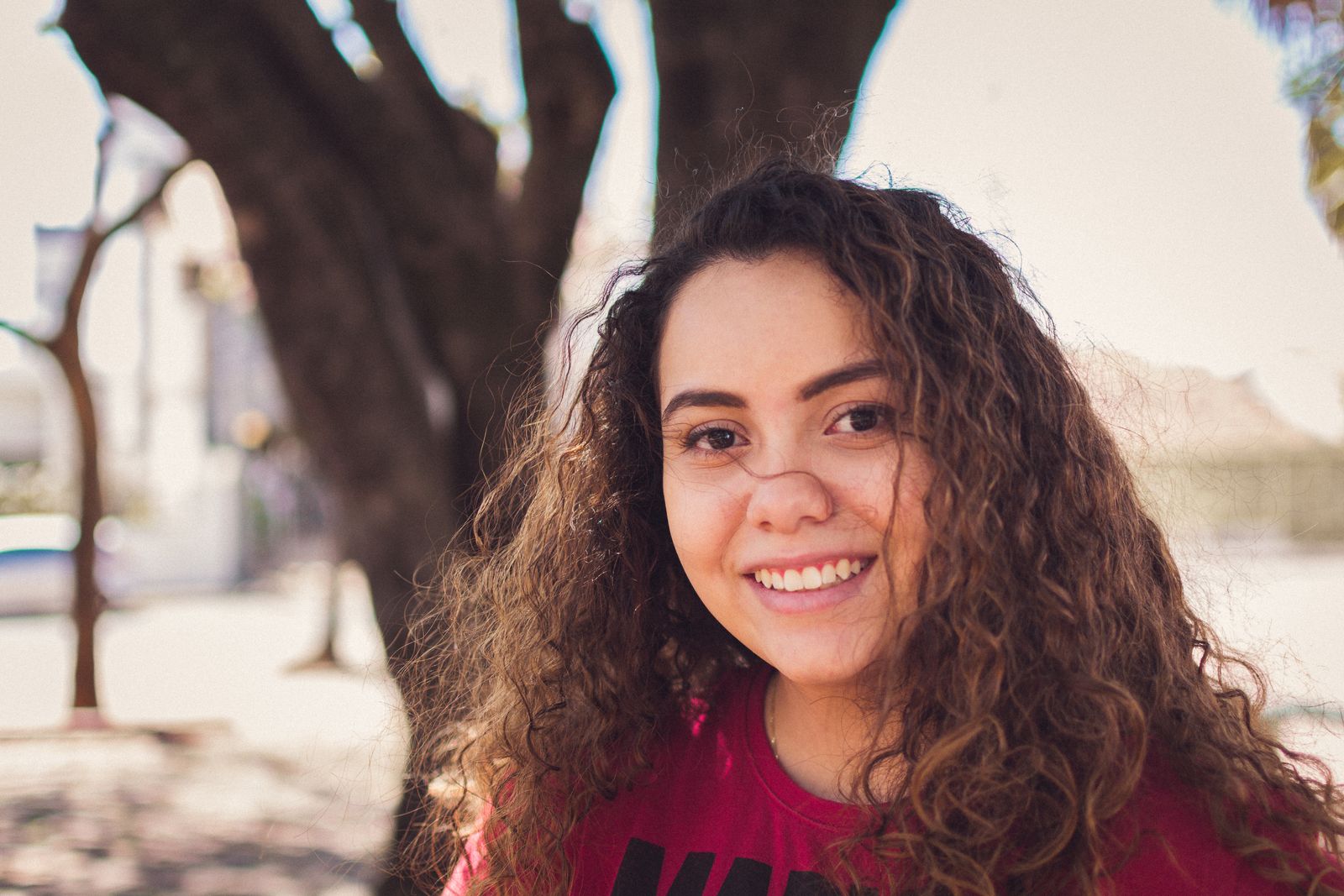 Portrait of a smiling young woman with curly hair in a sunny outdoor setting.