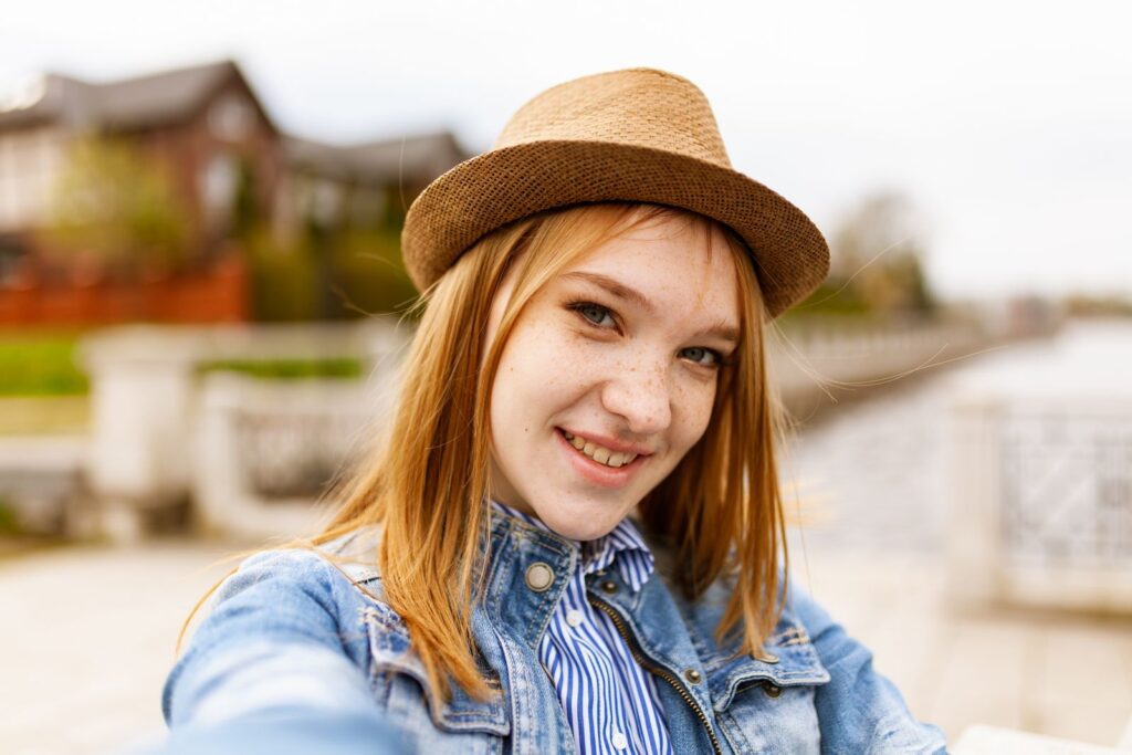 Cheerful young woman wearing a denim jacket and fedora hat smiling outdoors.