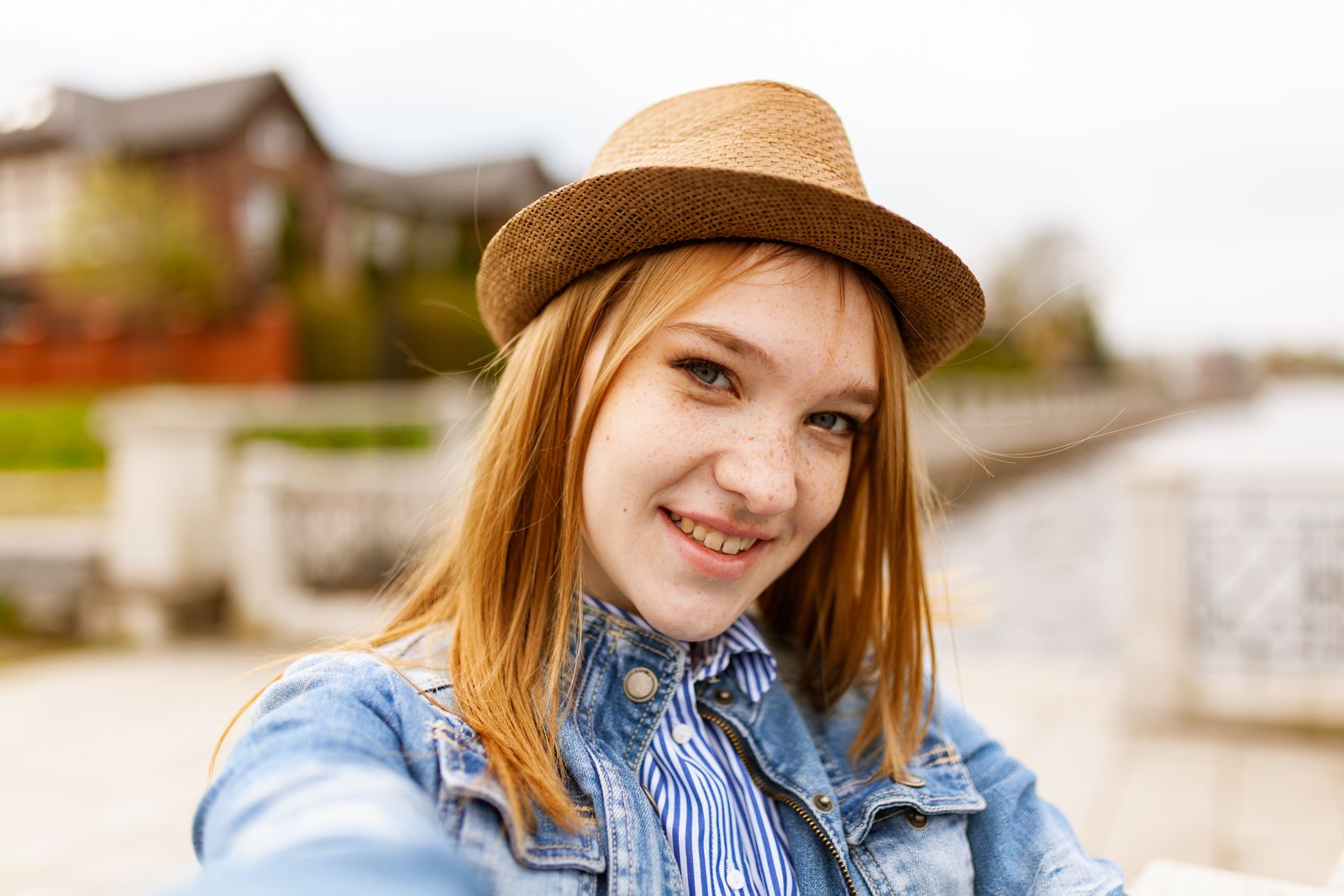 Cheerful young woman wearing a denim jacket and fedora hat smiling outdoors.
