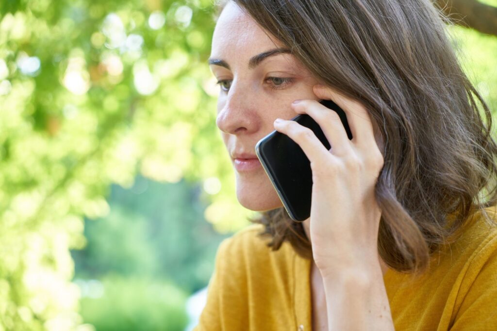 Side profile of a woman using a smartphone outdoors in a sunlit park, wearing a yellow top.