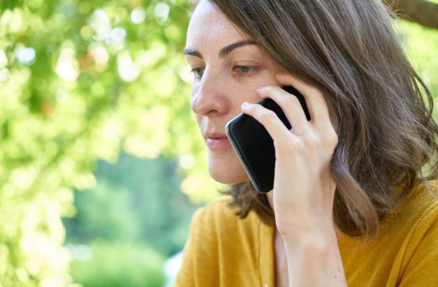 Side profile of a woman using a smartphone outdoors in a sunlit park, wearing a yellow top.