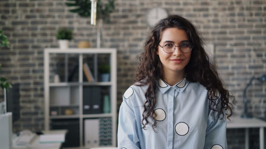 Smiling young woman in stylish office setting, wearing eyeglasses and patterned shirt.