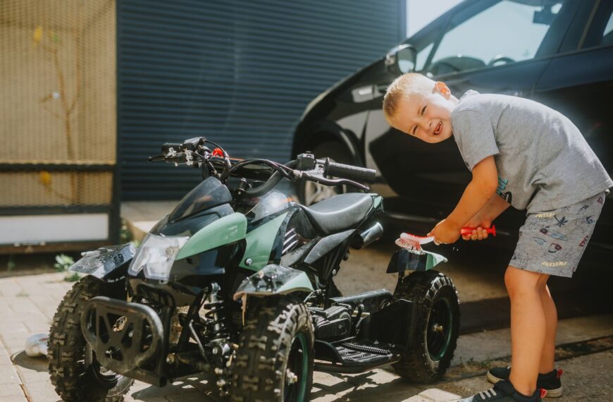 A young boy cheerfully cleans a kids' ATV outdoors on a sunny day, showcasing a playful moment.