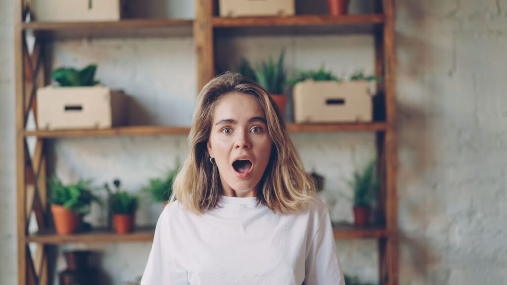 Young woman in white shirt with a surprised expression indoors, surrounded by plants.