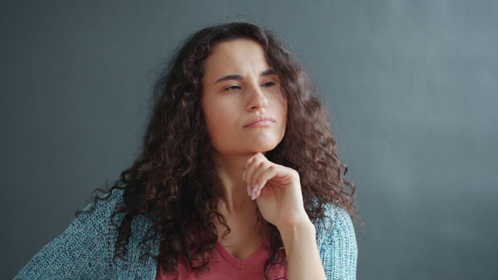 A young woman with curly hair ponders deeply, hand on chin against a gray backdrop.