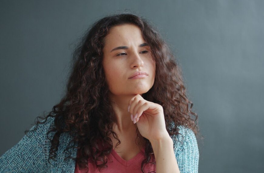 A young woman with curly hair ponders deeply, hand on chin against a gray backdrop.