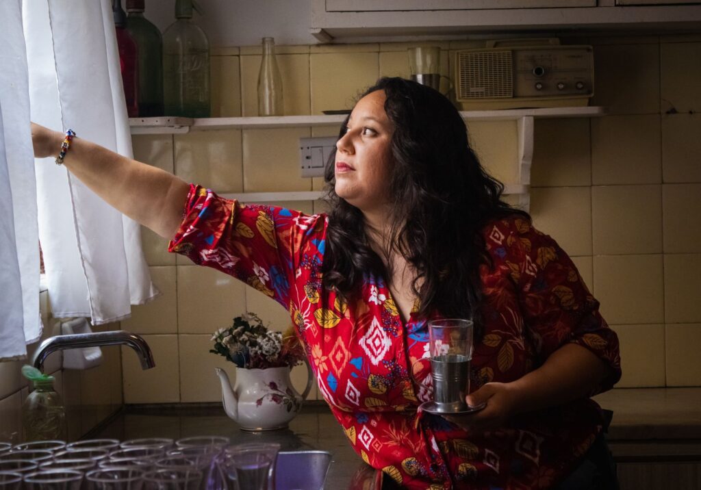 A woman in a colorful shirt adjusts a curtain in a cozy kitchen, holding a glass.