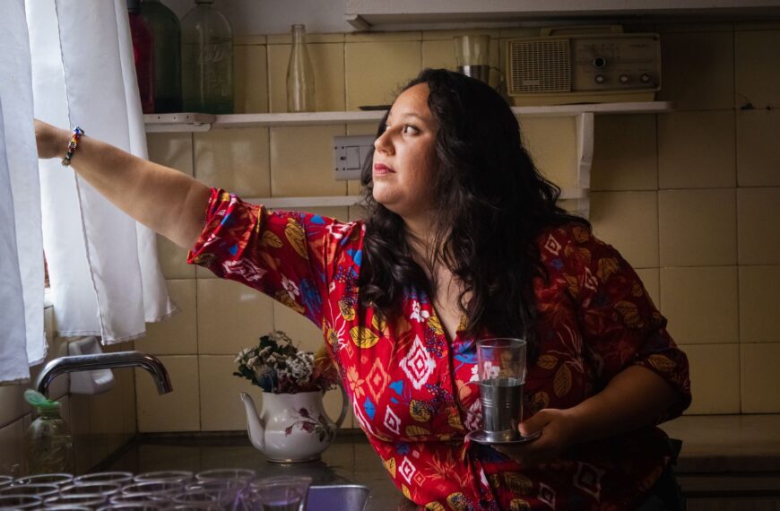 A woman in a colorful shirt adjusts a curtain in a cozy kitchen, holding a glass.