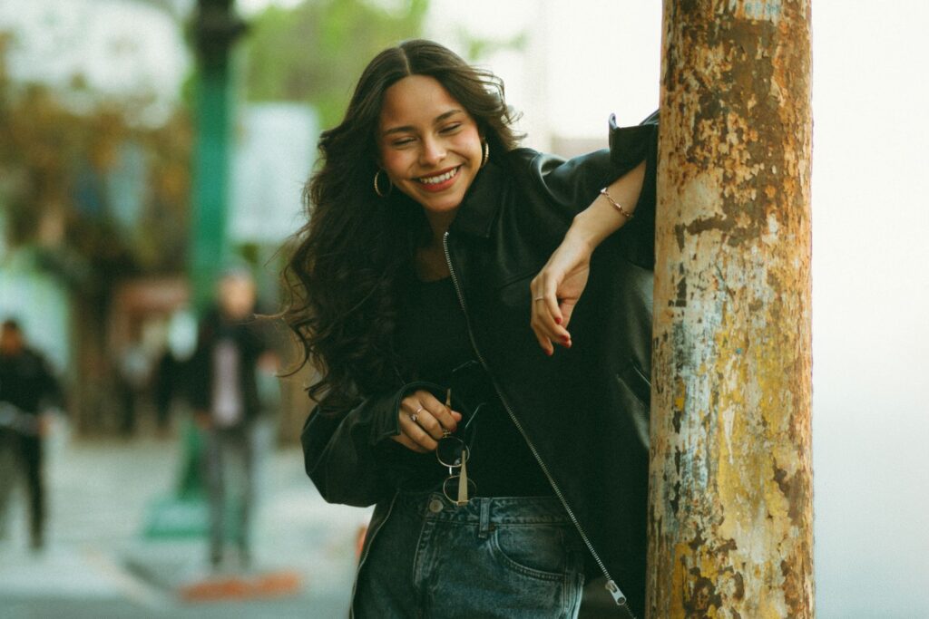 Smiling young woman in casual outfit leaning on a pole in urban setting. Vibrant and candid street portrait.