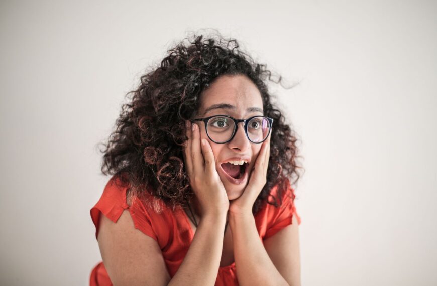 A woman expresses surprise, her curly hair framing her eyeglass-wearing face, hands on cheeks.