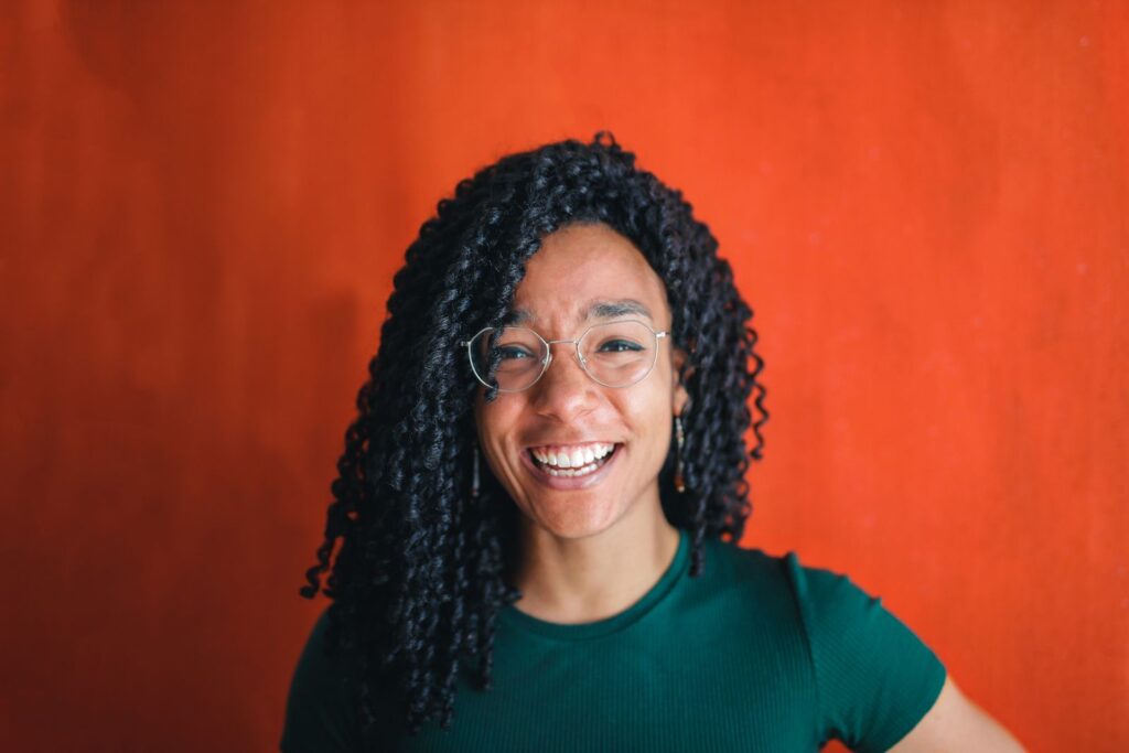 A joyful young woman with curly hair and glasses, smiling brightly against a vibrant red backdrop.