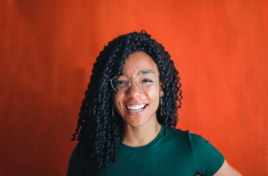 A joyful young woman with curly hair and glasses, smiling brightly against a vibrant red backdrop.