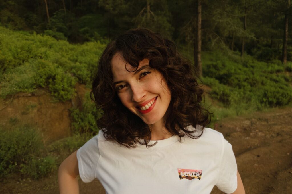 A cheerful woman with curly hair smiling outdoors in a green forest setting.