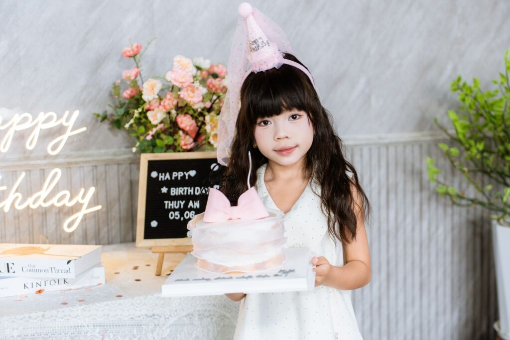 Adorable girl in a party hat holding a birthday cake surrounded by floral decorations.