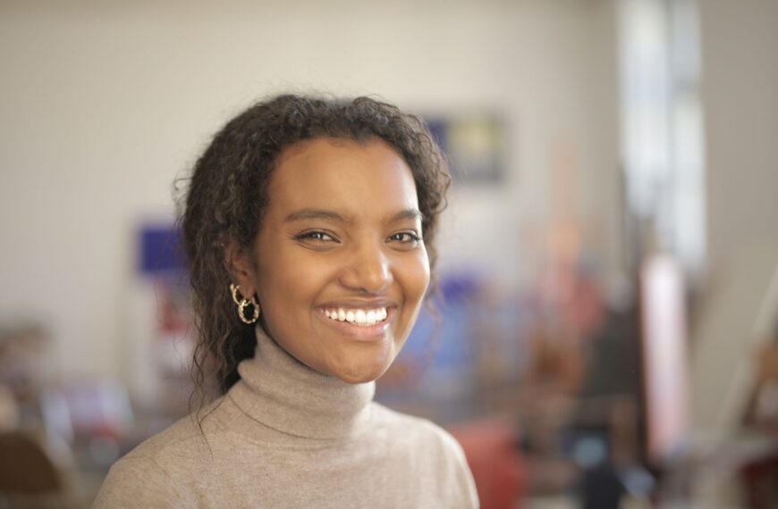 A cheerful woman with curly hair smiling indoors, wearing a turtleneck.