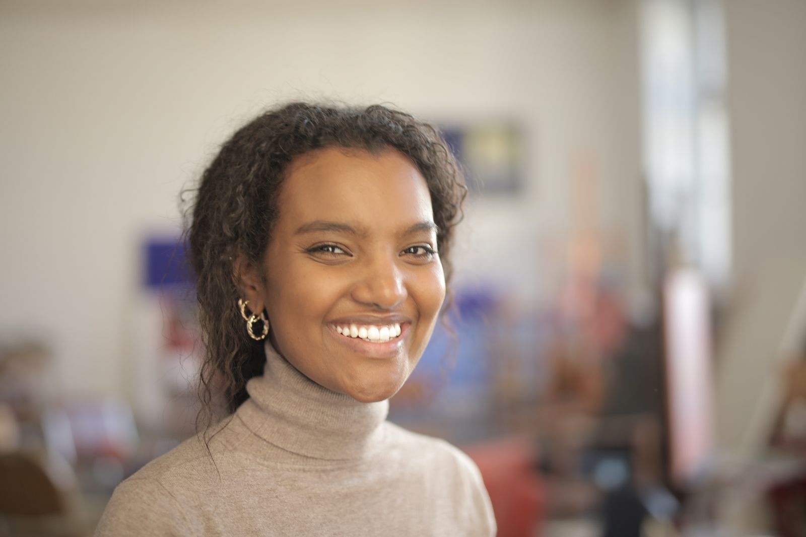 A cheerful woman with curly hair smiling indoors, wearing a turtleneck.