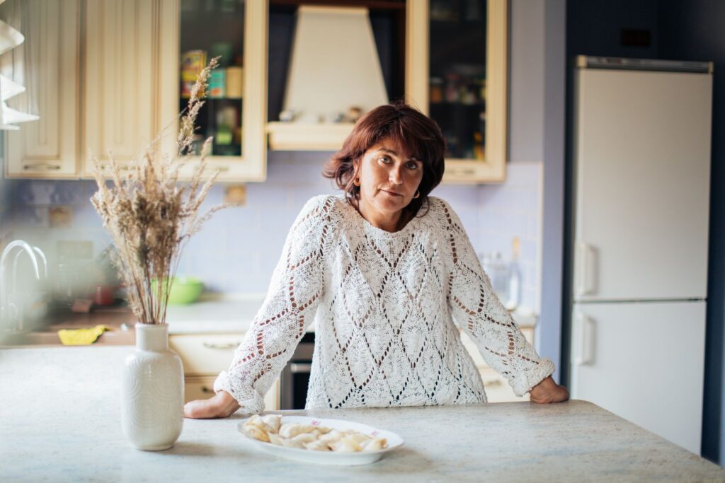 Woman in a kitchen with pelmeni on counter, wearing knitted sweater.