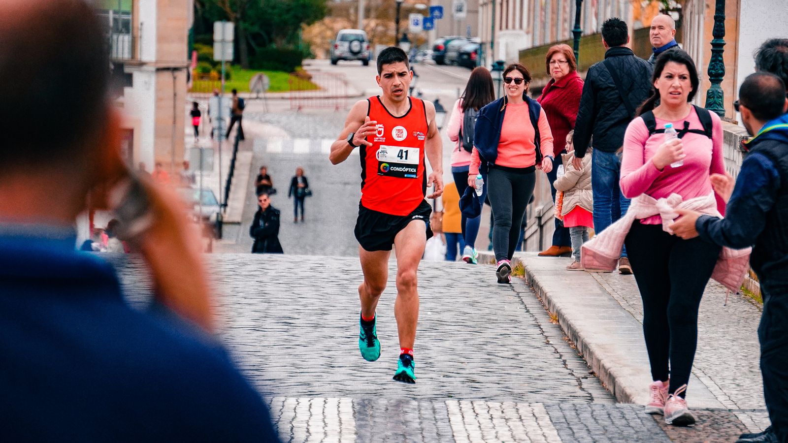 A male athlete runs a marathon through a bustling urban area on a cobblestone street.