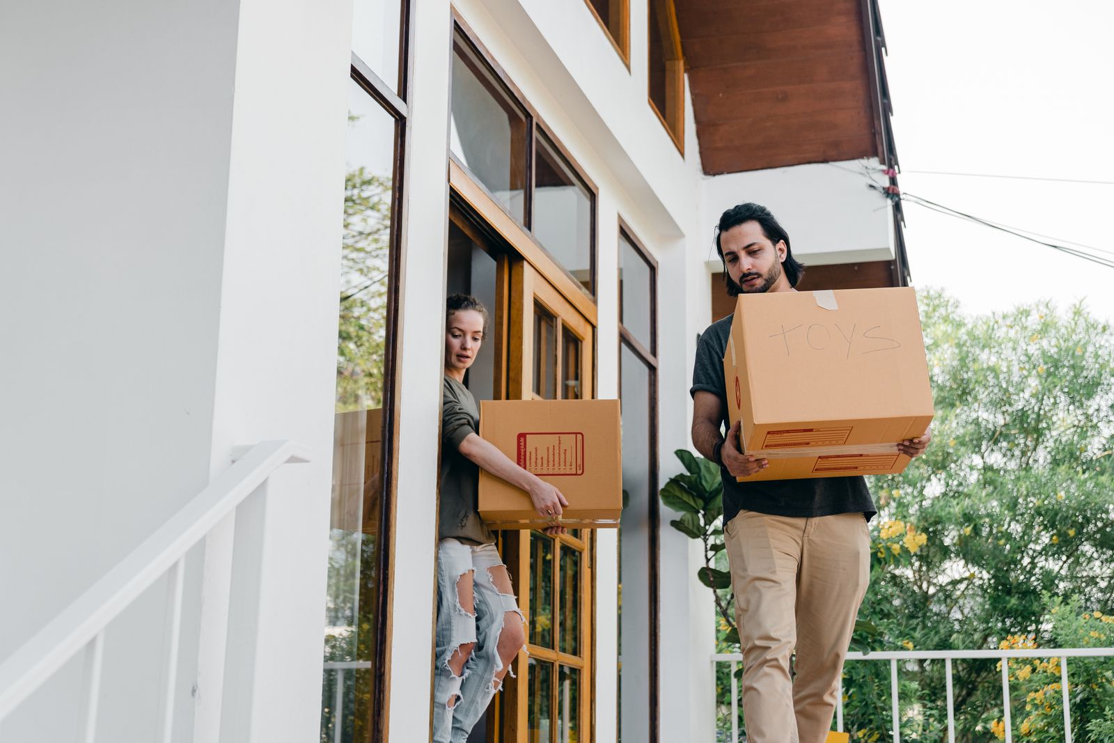 Young diverse male and female in modern casual outfits with cardboard boxes in hands watching step while moving out of old house on summer day