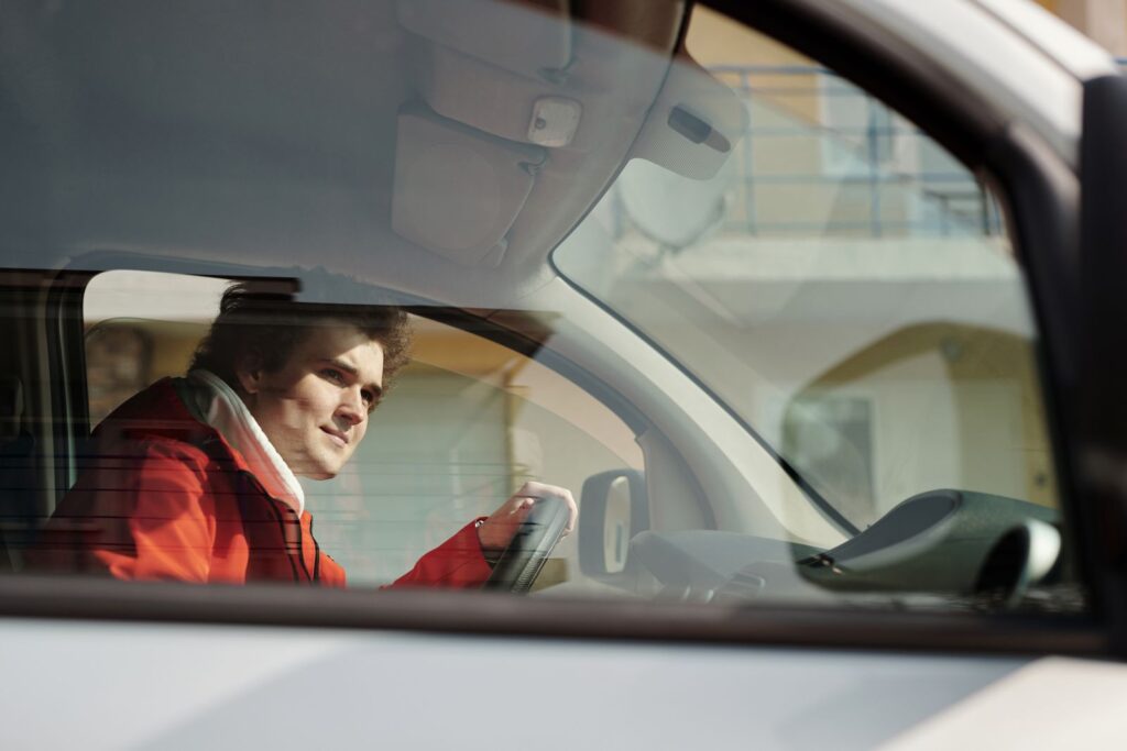 A man in a red jacket driving a car, seen through the window on a sunny day.