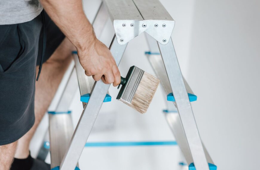 Side view of crop anonymous male master standing on ladder with tool while painting walls in flat