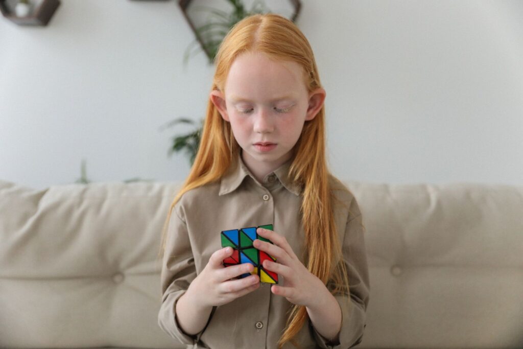 Adorable clever girl with red hair pondering over difficult colorful brain teaser cube