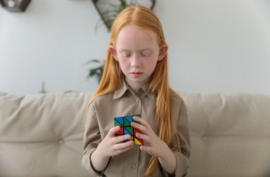 Adorable clever girl with red hair pondering over difficult colorful brain teaser cube