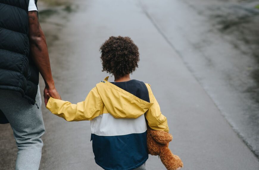 From above back view of crop anonymous ethnic father holding boy with toy bear by hand on asphalt roadway