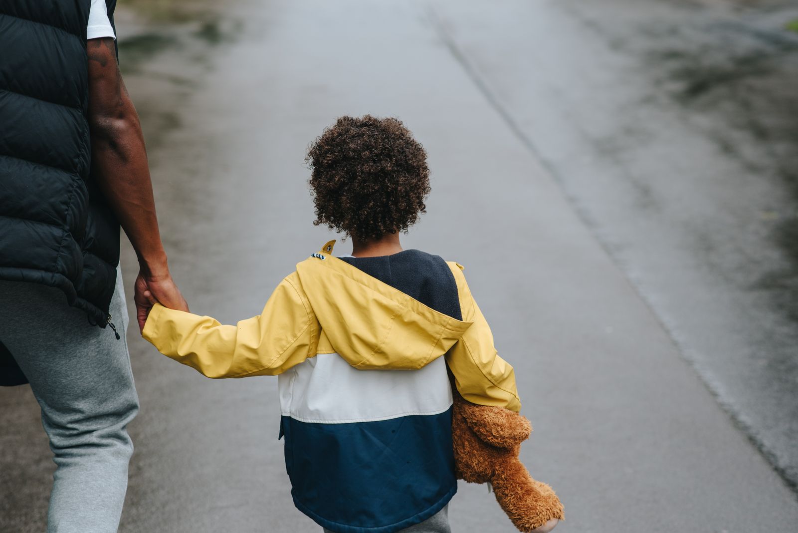 From above back view of crop anonymous ethnic father holding boy with toy bear by hand on asphalt roadway