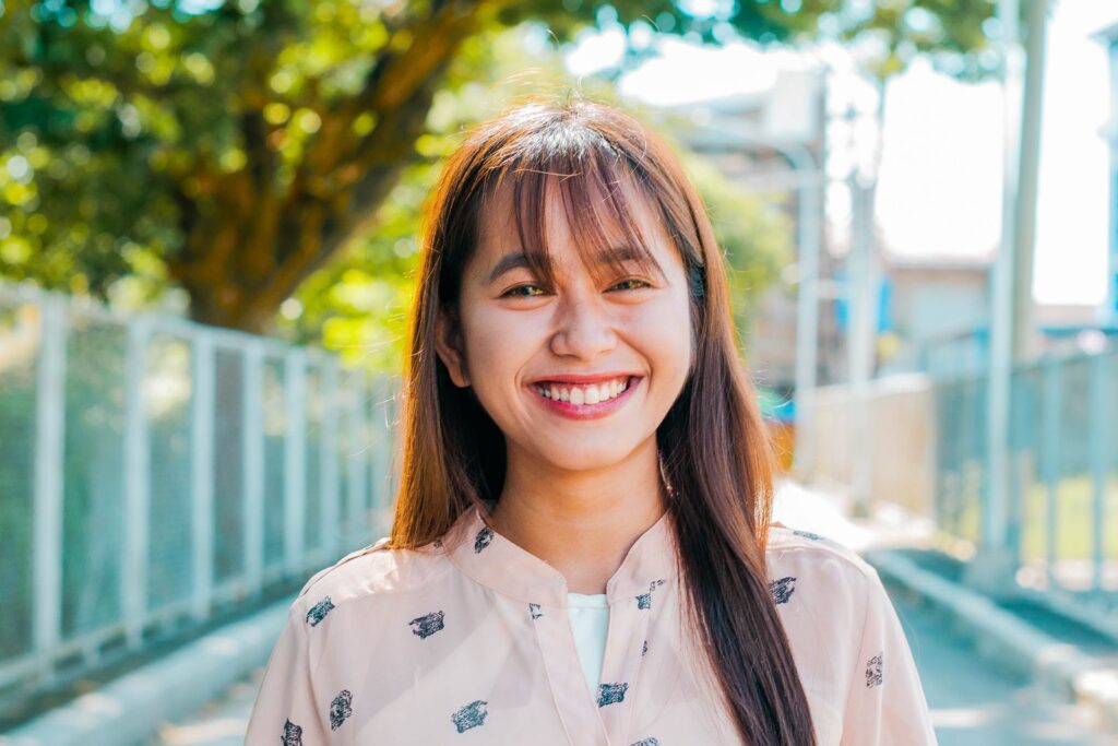 Positive Asian female with long hair looking at camera with smile while standing on sunny street with trees and metal fences on blurred background